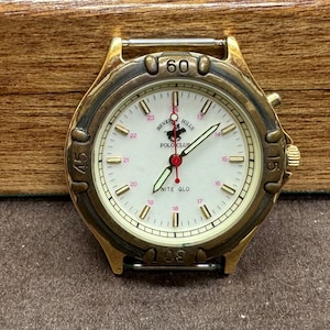 May include: A gold-toned wristwatch with a white face and black hands. The watch face has the words "Beverly Hills Polo Club" and "Nite Glo" printed on it. The watch has a black band.