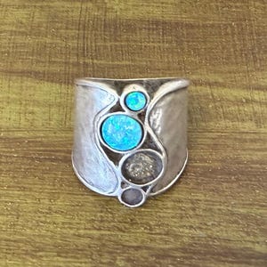 May include: A wide silver-toned ring featuring a textured, matte finish. The center of the band is adorned with four circular inlays of varying sizes, including two bright blue iridescent stones and two dark, neutral-toned circular accents.