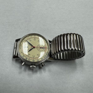 May include: A vintage-style wristwatch with a cream-colored face, black numerals, and silver hands. The watch has a silver case and a silver expanding band. The watch face includes multiple sub-dials and a red second hand. The watch is displayed on a white surface.