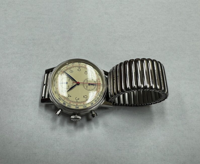 May include: A vintage-style wristwatch with a cream-colored face, black numerals, and silver hands. The watch has a silver case and a silver expanding band. The watch face includes multiple sub-dials and a red second hand. The watch is displayed on a white surface.
