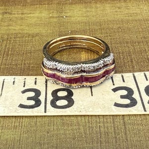 May include: A gold ring with a layered design. The ring features a row of small, clear stones, a row of red stones, and a gold band. The ring is on a measuring tape.