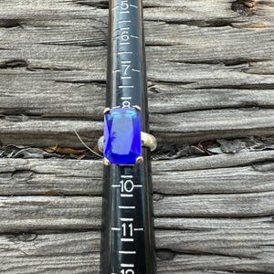 May include: A silver ring with a large, rectangular, deep blue gemstone. The ring is set on a black ring sizer, with measurements in inches. The background is weathered wood.