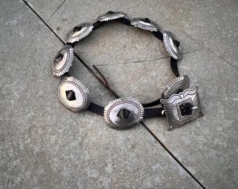 May include: A silver belt with multiple decorative conchos. The conchos are oval shaped with a black center and intricate designs around the edges. The belt is made of black leather.
