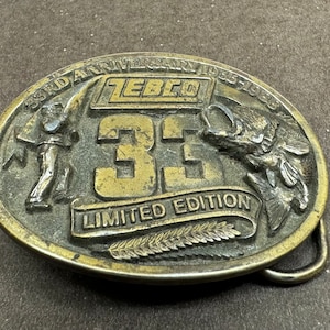 May include: Oval-shaped, brass-colored belt buckle. The buckle features the text "33RD ANNIVERSARY 1955-1988" and "ZEBCO" above the large number "33". Below the number is the text "LIMITED EDITION". The buckle has a fishing theme.