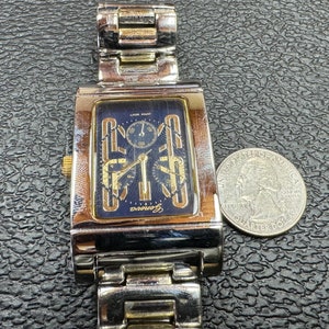 K&ouml;nnte beinhalten: Eine Herren-Armbanduhr in Silber und Gold mit schwarzem Zifferblatt und drei Chronographen-Zifferbl&auml;ttern. Die Uhr hat ein Edelstahlband und ein rechteckiges Geh&auml;use.