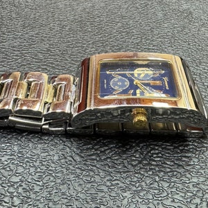 K&ouml;nnte beinhalten: Eine silberne und goldene Herrenarmbanduhr mit blauem Zifferblatt und einem silbernen Metallband. Die Uhr hat ein quadratisches Zifferblatt und einen Chronographen.