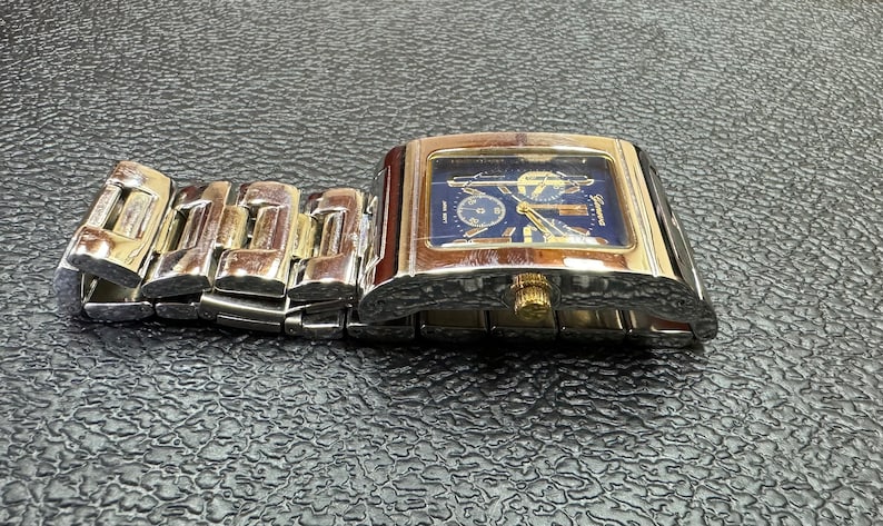 K&ouml;nnte beinhalten: Eine silberne und goldene Herrenarmbanduhr mit blauem Zifferblatt und einem silbernen Metallband. Die Uhr hat ein quadratisches Zifferblatt und einen Chronographen.
