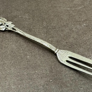 May include: A silver fork with a rose design on the handle. The fork has three tines and is ornate.