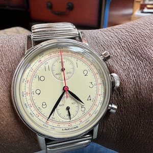 May include: A silver wristwatch with a cream-colored face, black hands, and a red second hand. The watch features a chronograph function and is marked with the brand name "UNDONE". The watch is on a silver metal band.