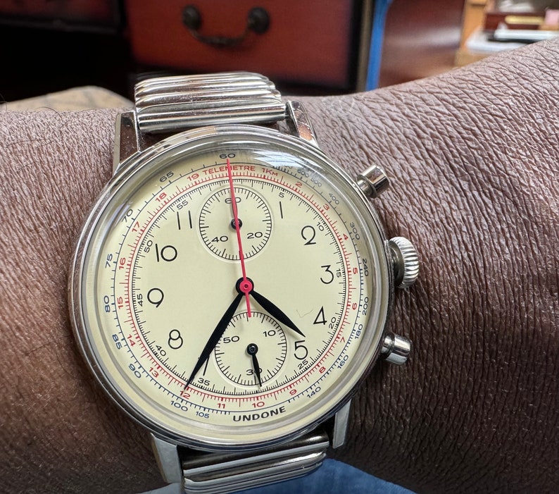 May include: A silver wristwatch with a cream-colored face, black hands, and a red second hand. The watch features a chronograph function and is marked with the brand name "UNDONE". The watch is on a silver metal band.