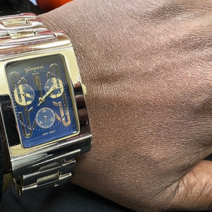 K&ouml;nnte beinhalten: Eine goldfarbene Uhr mit quadratischem Zifferblatt und blauem Zifferblatt. Die Uhr hat eine Chronographenfunktion und der Text "Geneva" ist auf dem Zifferblatt aufgedruckt.