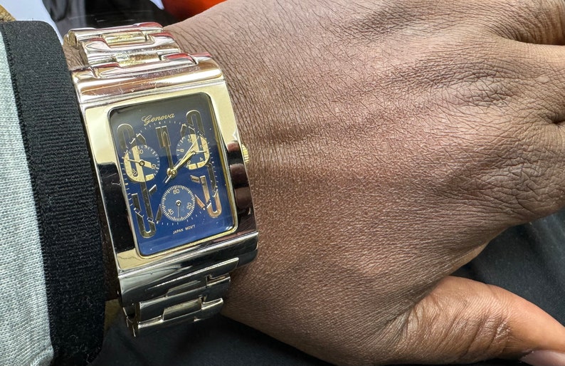 K&ouml;nnte beinhalten: Eine goldfarbene Uhr mit quadratischem Zifferblatt und blauem Zifferblatt. Die Uhr hat eine Chronographenfunktion und der Text "Geneva" ist auf dem Zifferblatt aufgedruckt.