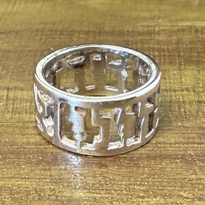 May include: A silver ring with a repeating cut-out pattern. The ring has a wide band with a series of stylized cross shapes. The ring is made of a shiny, metallic material and sits on a wooden surface.