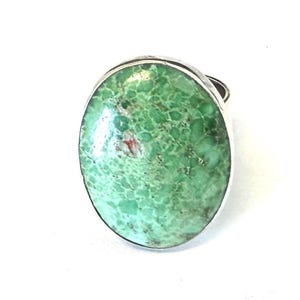 May include: Oval-shaped ring featuring a large, light green stone with a textured surface and a silver-colored band. The stone has a mottled appearance with hints of red.