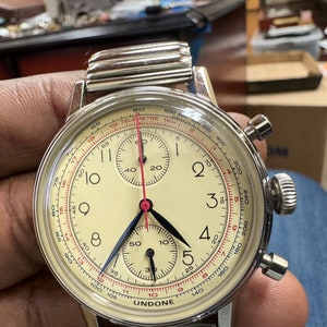 May include: A vintage-style UNDONE chronograph watch with a cream-colored face, black and blue hands, and red accents. The watch features multiple sub-dials and a stainless steel band. The watch has a telemetre scale and is approximately 40mm in diameter.