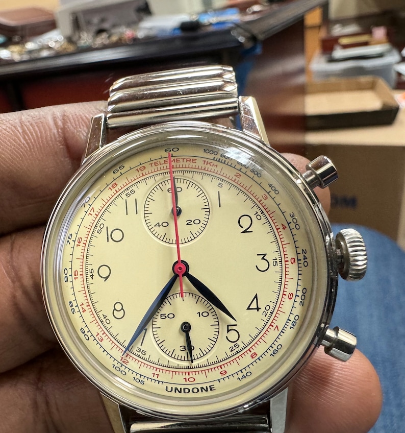 May include: A vintage-style UNDONE chronograph watch with a cream-colored face, black and blue hands, and red accents. The watch features multiple sub-dials and a stainless steel band. The watch has a telemetre scale and is approximately 40mm in diameter.