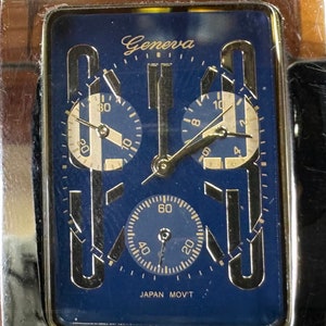 K&ouml;nnte beinhalten: Eine silberne und goldene Chronographen-Armbanduhr mit blauem Zifferblatt. Das Zifferblatt der Uhr hat drei Hilfszifferbl&auml;tter und den Text "Geneva" und "JAPAN MOV'T".