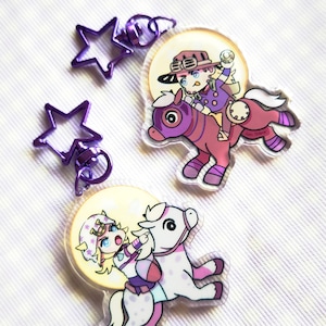 Puede incluir: Dos llaveros de acrílico con personajes estilo anime montando caballos. Un llavero muestra a un personaje con sombrero marrón sobre un caballo morado, el otro a un personaje con tocado blanco sobre un caballo blanco. Tienen cierres de estrella morados.