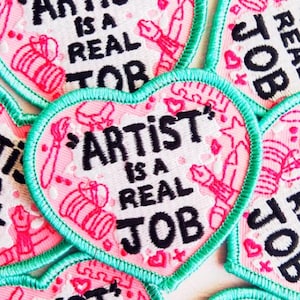 Puede incluir: Parches bordados en forma de corazón con el texto "ARTIST IS A REAL JOB" en negro. Los parches tienen un fondo rosa con ilustraciones de materiales de arte y un borde turquesa.