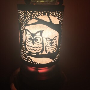 May include: A white ceramic oil diffuser with a black metal owl design. The diffuser has a glass top and a black base. The owls are silhouetted against a tree branch and a moon.