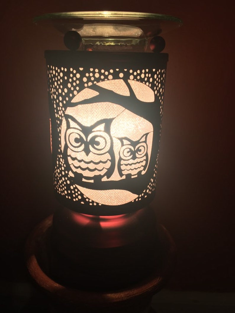 May include: A white ceramic oil diffuser with a black metal owl design. The diffuser has a glass top and a black base. The owls are silhouetted against a tree branch and a moon.