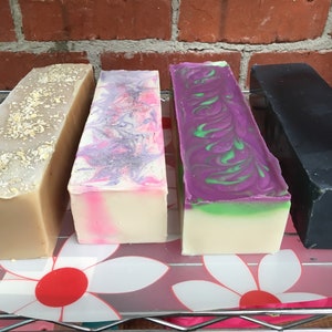 May include: Four bars of handmade soap, each with a different color and design. The bars are arranged in a row on a white and pink floral patterned surface. The soap bars are from left to right: beige with oatmeal, pink and purple swirl, green and purple swirl, and black.