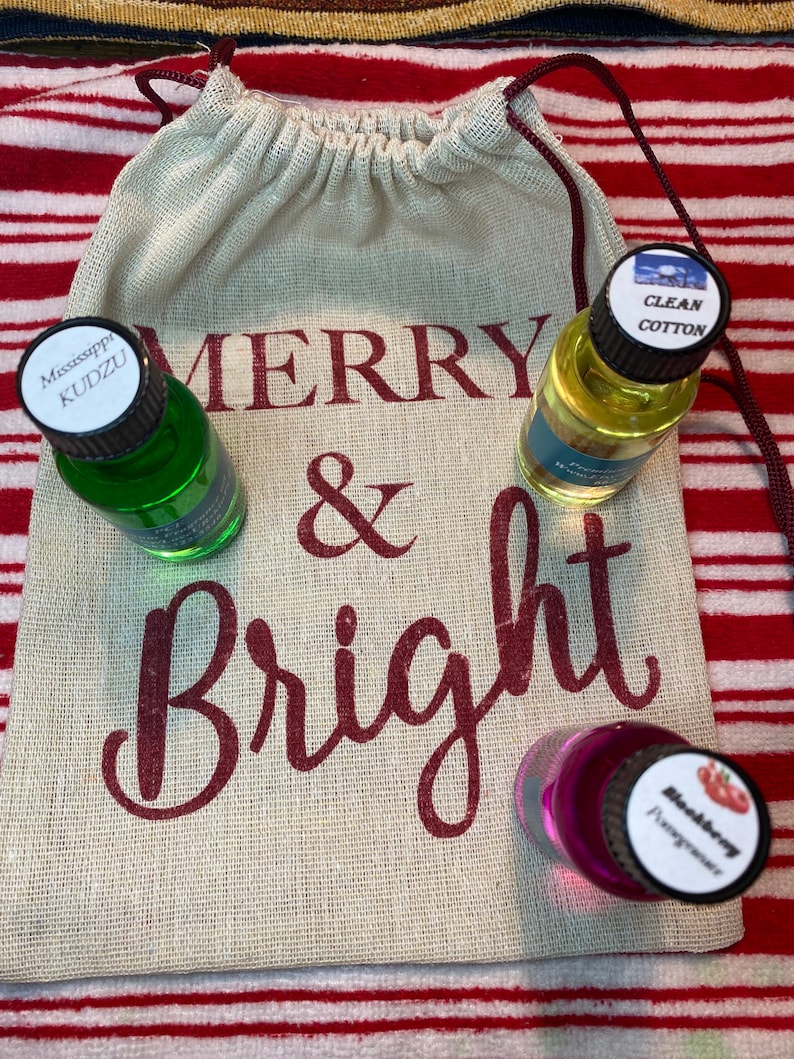May include: A burlap bag with the words "Merry & Bright" printed on it. Three small glass bottles with labels are inside the bag. The labels read "Mississippi Kudzu", "Clean Cotton", and "Blackberry Fragrance".