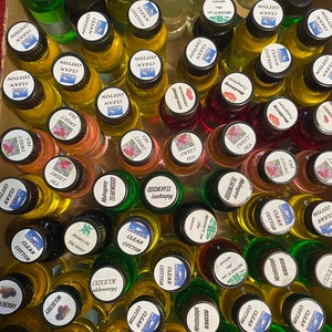 K&ouml;nnte beinhalten: Eine Sammlung kleiner Glasflaschen, gef&uuml;llt mit verschiedenfarbigen Duft&ouml;len, jeweils mit einem schwarzen Verschluss und einem wei&szlig;en, runden Etikett versehen. Die Etiketten tragen aufgedruckte Texte zur Identifizierung der D&uuml;fte.