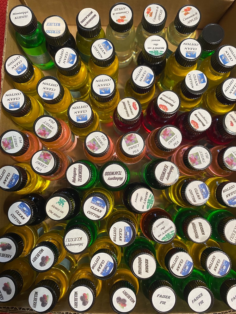 K&ouml;nnte beinhalten: Eine Sammlung kleiner Glasflaschen, gef&uuml;llt mit verschiedenfarbigen Duft&ouml;len, jeweils mit einem schwarzen Verschluss und einem wei&szlig;en, runden Etikett versehen. Die Etiketten tragen aufgedruckte Texte zur Identifizierung der D&uuml;fte.