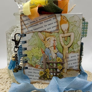 May include: A handmade journal featuring a Peter Rabbit illustration. The journal has a spiral binding, blue ribbon, and decorative embellishments. The cover art depicts Peter Rabbit with a carrot and a bird perched on a shovel. The journal is on a cream-colored crocheted surface.