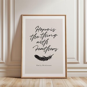 Puede incluir: Un cuadro enmarcado con la cita "Hope is the thing with feathers" en elegante escritura negra. Una ilustración de pluma negra está debajo de la cita, con "Emily Dickinson" debajo. El cuadro tiene un fondo neutro y un marco de madera.