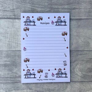 May include: White notepad with a lined interior and a border of cherries, cakes, and a dog wearing an apron. The notepad has the word "Recipes" printed at the top and the text "Hayley Walker Designs." at the bottom.