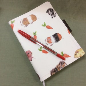 Peut inclure: Une couverture de carnet blanche avec un motif r&eacute;p&eacute;t&eacute; de cochons d'Inde et de carottes. Un stylo rouge est pos&eacute; sur la couverture.