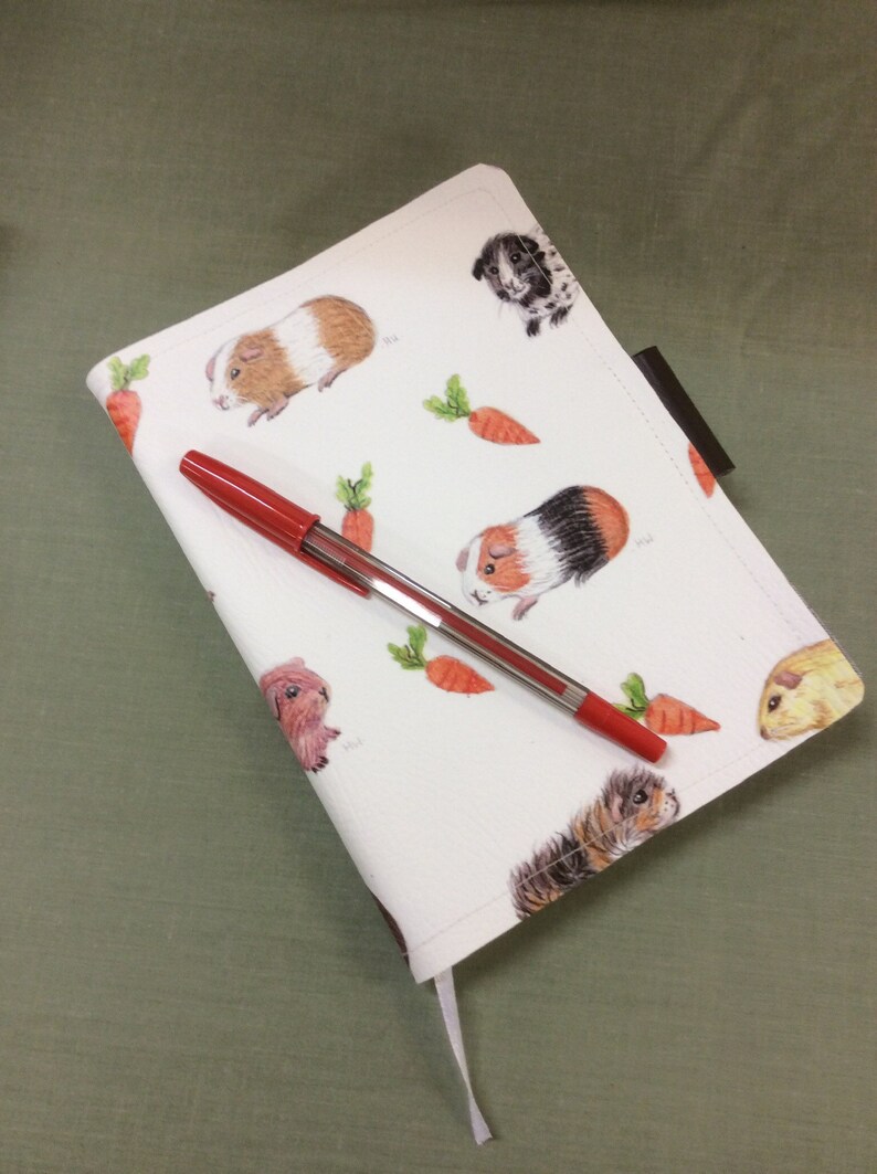 Peut inclure: Une couverture de carnet blanche avec un motif r&eacute;p&eacute;t&eacute; de cochons d'Inde et de carottes. Un stylo rouge est pos&eacute; sur la couverture.
