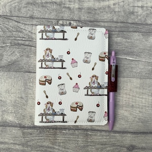 May include: A white notebook cover with a repeating pattern of guinea pigs baking. The pattern includes flour bags, spoons, cherries, and cupcakes. A purple pen is attached to the cover.