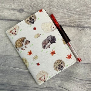 May include: A white leather notebook cover with a hedgehog pattern. The cover features hedgehogs of different colours and sizes, as well as red ladybirds and green leaves. A red pen is tucked into the cover.