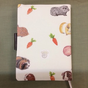 Peut inclure: Couverture de carnet blanche avec un motif r&eacute;p&eacute;t&eacute; de cochons d'Inde et de carottes. La couverture a un signet en cuir marron et un petit logo en forme de patte.