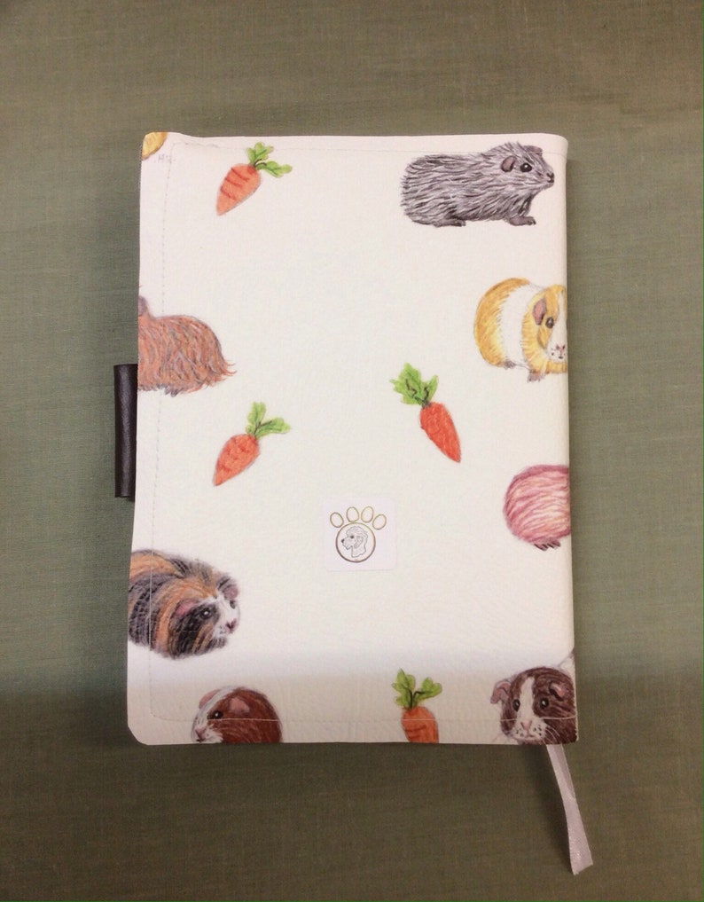Peut inclure: Couverture de carnet blanche avec un motif r&eacute;p&eacute;t&eacute; de cochons d'Inde et de carottes. La couverture a un signet en cuir marron et un petit logo en forme de patte.