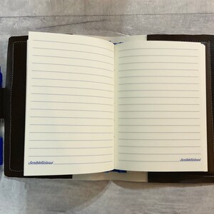 Peut inclure: Un journal reli&eacute; en cuir marron avec des pages lign&eacute;es et un porte-stylo. Le journal est ouvert sur une page blanche avec le mot "Scribblelicious" &eacute;crit en cursive en bas.