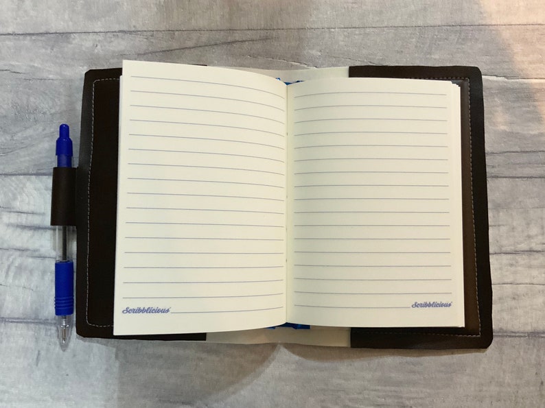 Peut inclure: Un journal reli&eacute; en cuir marron avec des pages lign&eacute;es et un porte-stylo. Le journal est ouvert sur une page blanche avec le mot "Scribblelicious" &eacute;crit en cursive en bas.