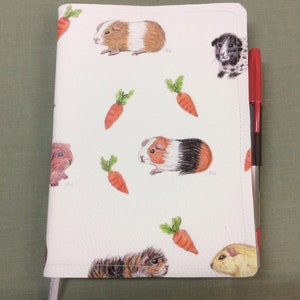 Peut inclure: Une couverture de carnet blanche avec un motif r&eacute;p&eacute;t&eacute; de cochons d'Inde et de carottes. La couverture a un ruban marque-page blanc et un stylo rouge et noir est gliss&eacute; dans la couverture.