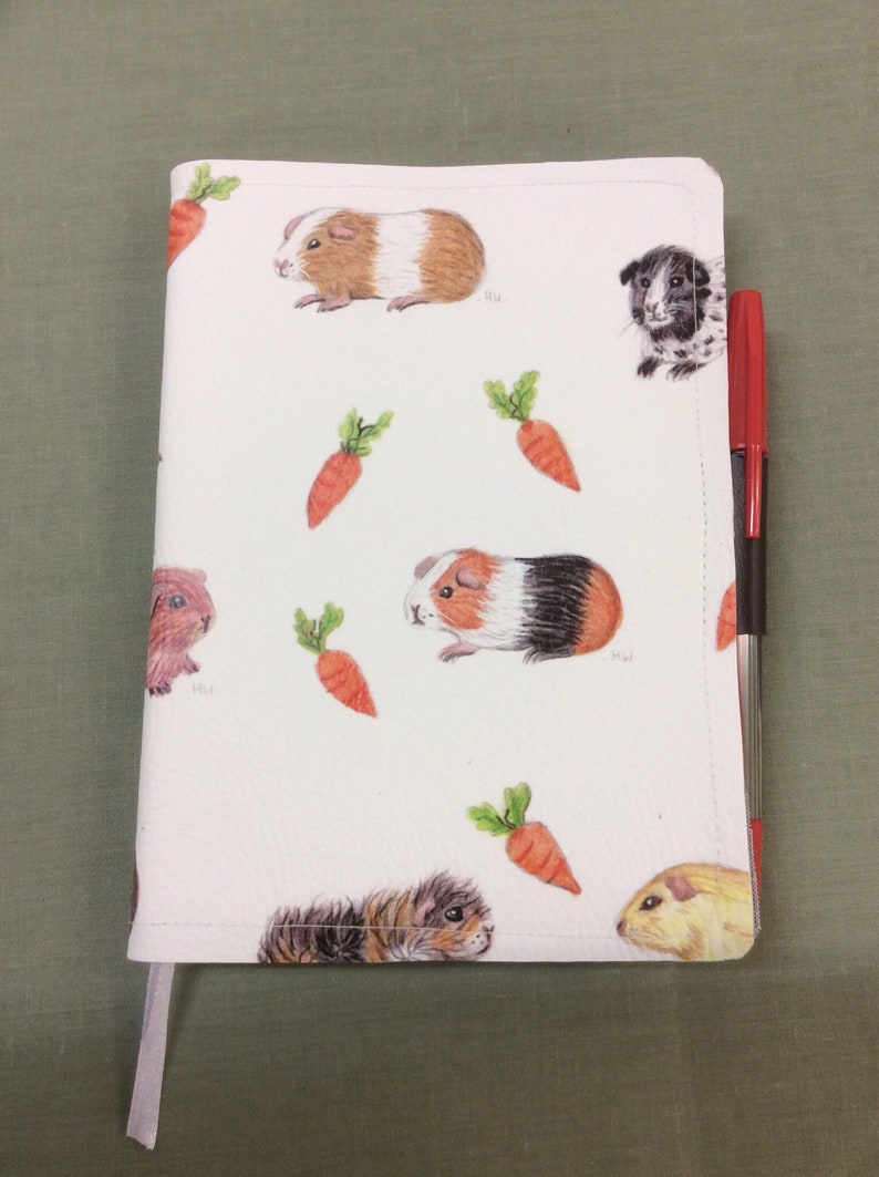 Peut inclure: Une couverture de carnet blanche avec un motif r&eacute;p&eacute;t&eacute; de cochons d'Inde et de carottes. La couverture a un ruban marque-page blanc et un stylo rouge et noir est gliss&eacute; dans la couverture.