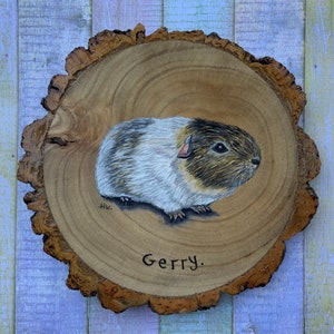 May include: A hand-painted wooden slice with a portrait of a guinea pig. The guinea pig is white with brown and black markings. The wood slice is brown and has a natural bark edge. The guinea pig is named "Gerry".