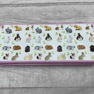 Puede incluir: Un estuche rectangular con borde rosa. El estuche está decorado con un estampado de varios conejos de diferentes colores y poses, junto con dientes de león amarillos sobre un fondo blanco.