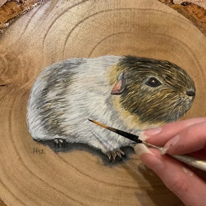 May include: A hand-painted portrait of a guinea pig on a wooden slice. The guinea pig is white with brown and black markings. The artist is using a small paintbrush to add details to the painting.
