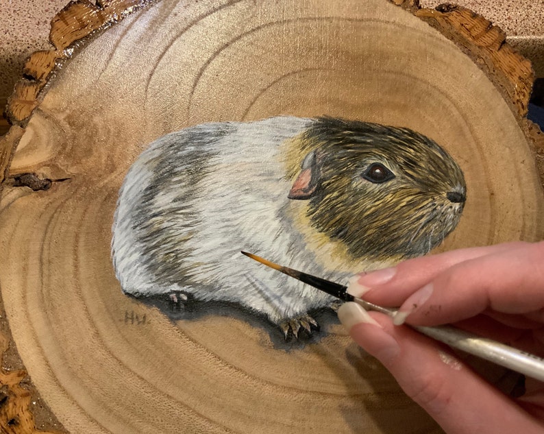 May include: A hand-painted portrait of a guinea pig on a wooden slice. The guinea pig is white with brown and black markings. The artist is using a small paintbrush to add details to the painting.