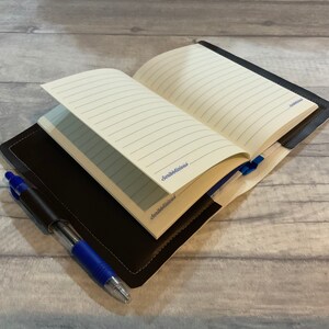 Peut inclure: Un journal reli&eacute; en cuir marron avec des pages lign&eacute;es, ouvert pour r&eacute;v&eacute;ler un stylo bleu gliss&eacute; dans la couverture.