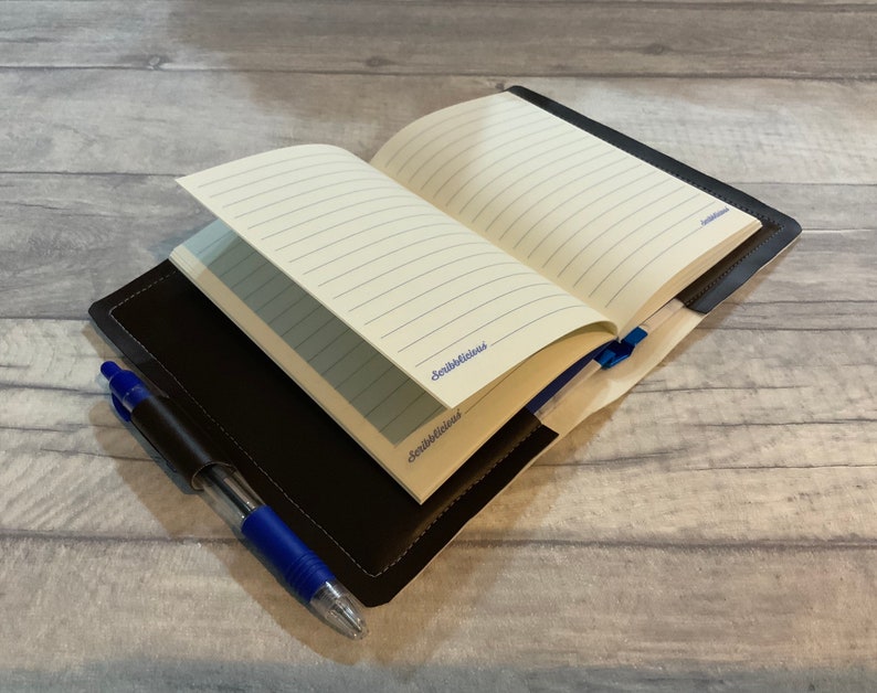 Peut inclure: Un journal reli&eacute; en cuir marron avec des pages lign&eacute;es, ouvert pour r&eacute;v&eacute;ler un stylo bleu gliss&eacute; dans la couverture.