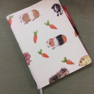 Peut inclure: Une couverture de carnet blanche avec un motif r&eacute;p&eacute;t&eacute; de cochons d'Inde et de carottes. La couverture est en mati&egrave;re textur&eacute;e et poss&egrave;de un marque-page en ruban blanc.