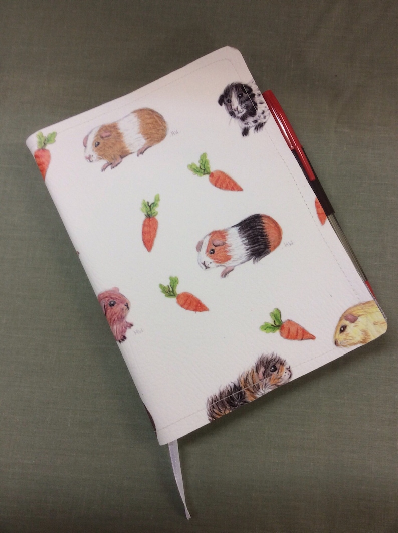 Peut inclure: Une couverture de carnet blanche avec un motif r&eacute;p&eacute;t&eacute; de cochons d'Inde et de carottes. La couverture est en mati&egrave;re textur&eacute;e et poss&egrave;de un marque-page en ruban blanc.
